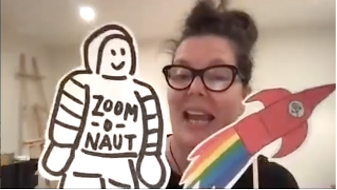 Screen capture from Workshop 1 video prompt showing a woman in glasses holding up a Zoom-o-Naut - a black and white hand-drawn astronaut in a space suit- and a red rocket with a rainbow coming out the bottom. 